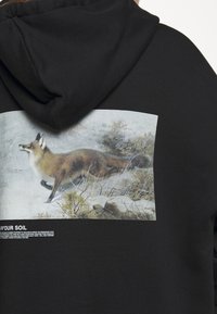 Sweat à capuche noir avec une image imprimée d'un renard marchant à travers des buissons enneigés dans le dos.
