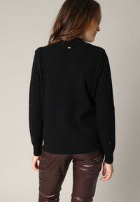 Vrouw met lang bruin haar, gekleed in een zwarte trui en donkerbruine leren broeken, met de rug naar de camera gekeerd tegen een effen lichte achtergrond.