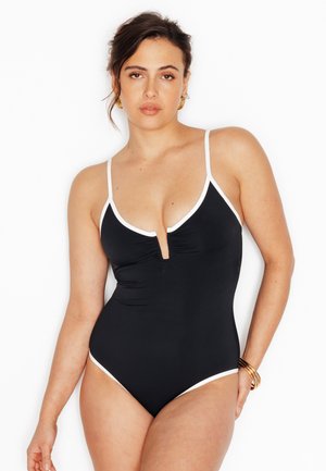 Donna che posa indossando un costume intero nero con rifiniture bianche e scollatura a V profonda, con orecchini a cerchio dorati e braccialetti.