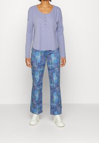 Lila långärmad topp med knappdetalj, matchad med blå mönstrade utsvängda byxor som visar abstrakta grafiker och vita sneakers.