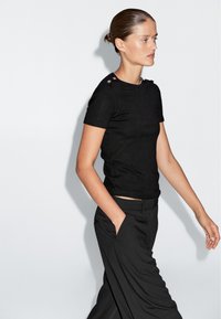 Zwarte getailleerde shirt met korte mouwen en knopenaccenten op de schouders, gecombineerd met losse zwarte broek, voor een effen witte achtergrond.