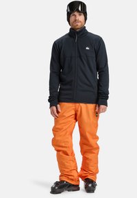 Čierna fleecová bunda na zips, oranžové lyžiarske nohavice a tmavé lyžiarske topánky. Bunda má logo na hrudi a otvory na palce na rukávoch.