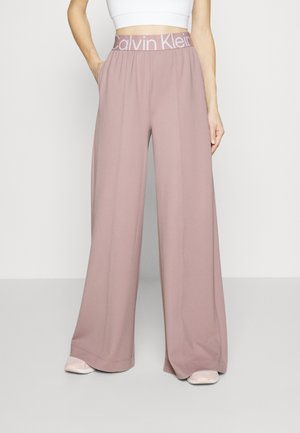 Mujer vistiendo pantalones anchos de color malva con cinturilla elástica con la marca Calvin Klein y una camiseta blanca, de pie con una mano en el bolsillo.