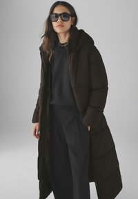 Manteau long et bouffi de couleur brune avec une capuche, fabriqué dans un matériau lisse. Associé à un haut sombre et un pantalon noir ample, complété par de grandes lunettes de soleil.