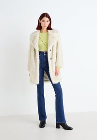 Femme aux cheveux rouges raides portant un manteau en fausse fourrure crème, un crop top torsadé vert citron, un jean bleu taille haute et des bottes à talons noires.