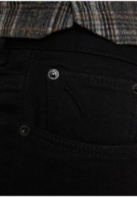 Jeans de mezclilla negros con un bolsillo visible y un pequeño detalle de logo bordado. Cuenta con botones de metal negro y costuras estructuradas.