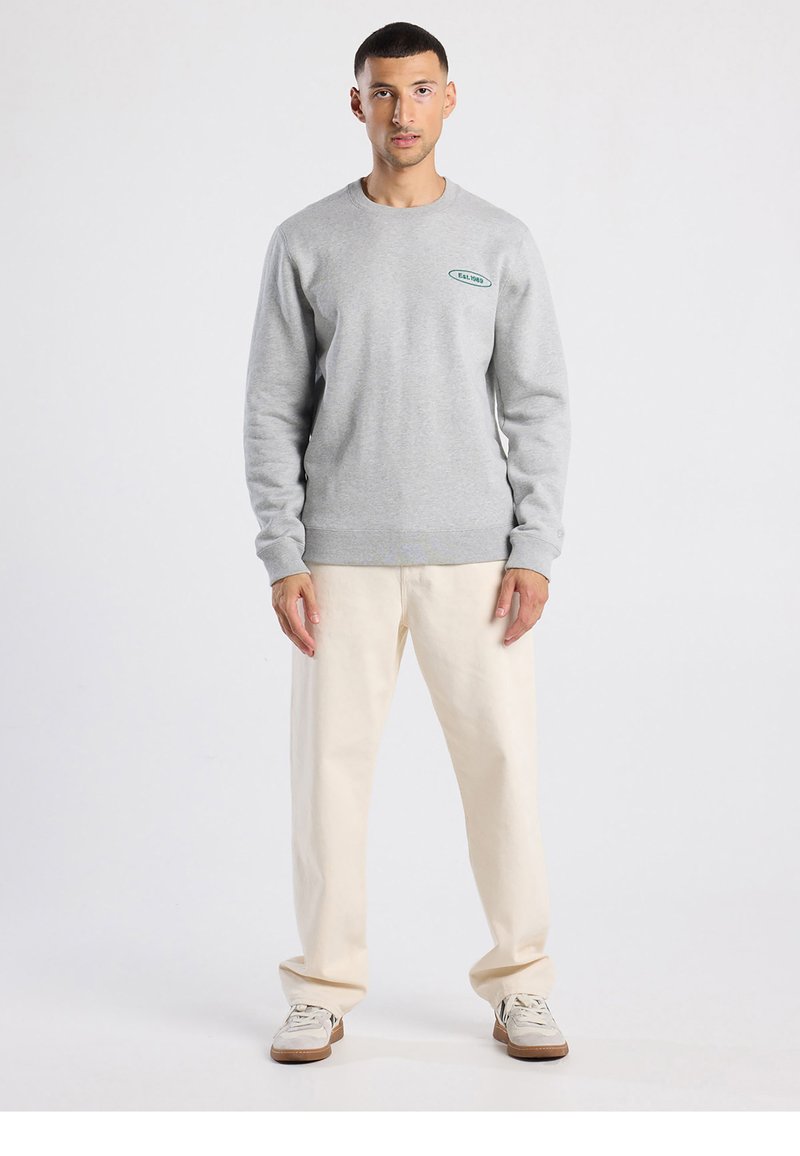 Grijze trui met ronde hals en geribbelde manchetten. Voorzien van een klein groen logo. Gecombineerd met crèmekleurige broek en lichte sneakers.