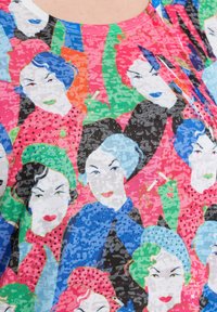 Farverig T-shirt med et gentaget mønster af illustrerede ansigtstræk med varierende frisurer og makeup. Materialet ser let og tekstureret ud.