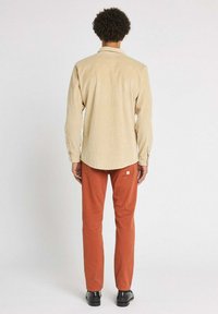 Chemise en velours côtelé beige avec col et poignets boutonnés, associée à un pantalon droit couleur rouille. Des chaussures noires complètent la tenue.