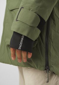Giacca esterna verde con tessuto testurizzato, polsino in neoprene nero con branding bianco e una fascia in velcro nera sulla manica.