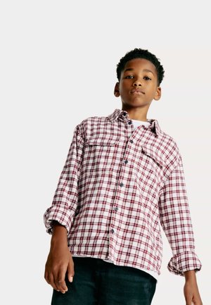 Ragazzo con capelli corti e ricci che indossa una camicia a quadri bianca e rossa con bottoni e pantaloni neri, che guarda con sicurezza davanti a sé su uno sfondo chiaro.