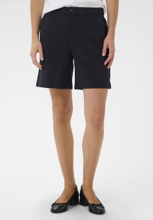 Personne portant un short noir sur mesure avec bouton et fermeture éclair, un haut blanc et des chaussures noires à talons bas, debout sur un fond blanc.