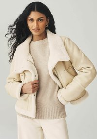 Vrouw met lang krullend zwart haar, gekleed in een beige schapenvachtjack over een beige trui en crèmekleurige broek, poserend tegen een effen lichte achtergrond.