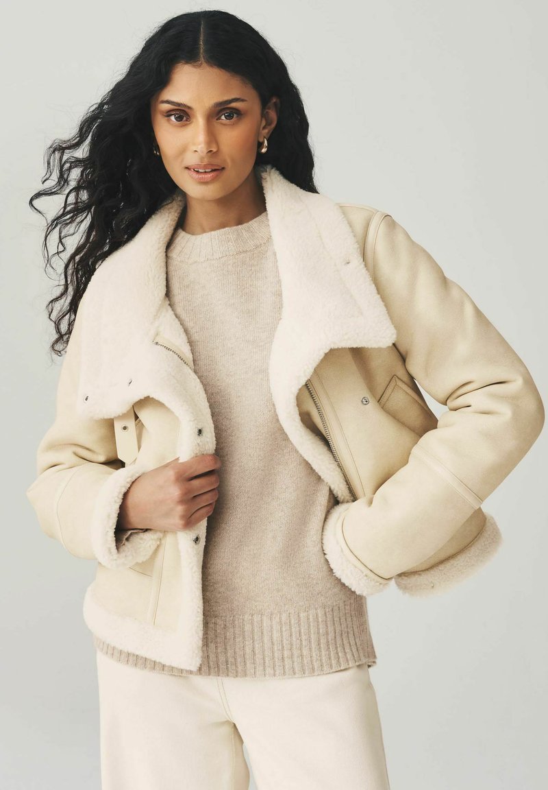 Vrouw met lang krullend zwart haar, gekleed in een beige schapenvachtjack over een beige trui en crèmekleurige broek, poserend tegen een effen lichte achtergrond.