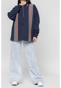 Sudadera navy con cremallera de un cuarto, que presenta rayas verticales en beige y rojo. Combinada con jeans de pierna ancha de color azul claro y zapatillas blancas.