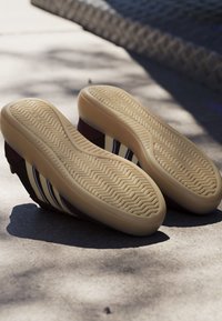 Paire de baskets avec semelles en gomme beige et côtés bordeaux avec des rayures blanches et bleues sur une surface en béton ensoleillée.