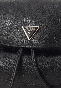 Bolso negro texturizado hecho de cuero sintético, con patrones en relieve y un logo triangular de Guess con acentos dorados.