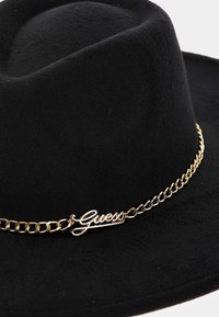 El sombrero de fieltro negro presenta una corona redondeada y una cadena dorada con un acento del logo "Guess". La textura es suave con un acabado liso.