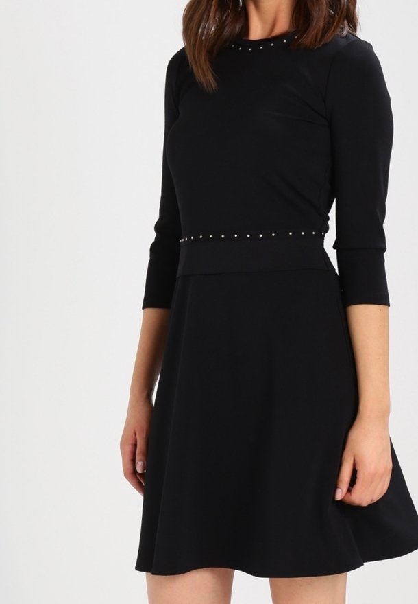 Femme portant une robe noire mi-longue avec des manches trois-quarts et de petites clous argentés le long de l'encolure et de la taille.