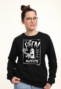 Felpa nera a maniche lunghe con una grande grafica bianca che raffigura un alieno in stile cartone animato e il testo "ALIEN INVASION".
