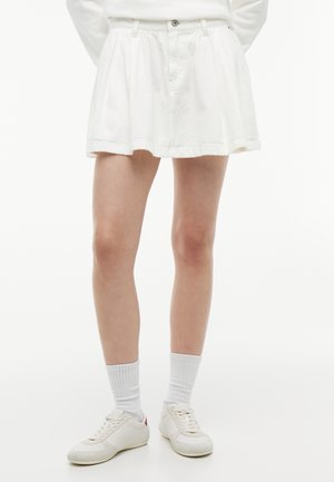 Personne portant une mini-jupe plissée blanche, des chaussettes blanches et des baskets blanches, debout devant un fond blanc uni.