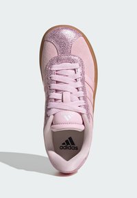 Pink athletic shoe with a textured upper, glittery accents, and three white stripes. Features flat laces and a rubber outsole.