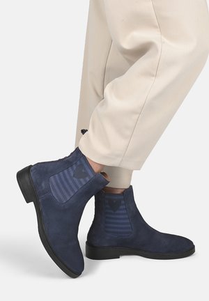 Navy blue suede Chelsea boots with striped elastic panels worn with beige pants on a white background.