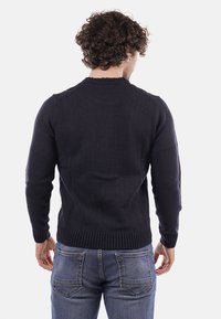 Uomo con capelli ricci che indossa un maglione nero lavorato a maglia e jeans blu, visto da dietro su uno sfondo uniforme.