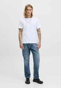 Vit T-shirt med rund hals, blå denimjeans och svarta snörskor. Modellen har tatueringar på armarna och ett silverhalsband.