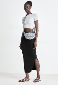 Donna che indossa un top grigio corto, una gonna nera con spacco alto, sandali bianchi e una borsa a tracolla metallica argento, in piedi davanti a uno sfondo bianco semplice.