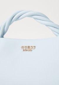 Borsa a mano blu chiaro in ecopelle testurizzata, con manici attorcigliati e un accento in metallo dorato con il logo che recita "GUESS EST. 1981."