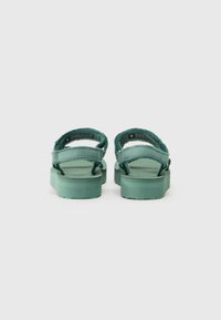 Green sandals with two adjustable straps, a textured rubber sole, and a flat design. Rear view highlights the strap placement and material.