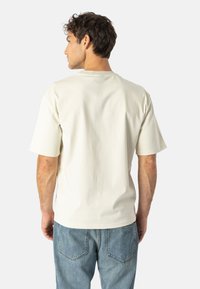 T-shirt in cotone crema a maniche corte con vestibilità rilassata e colletto rotondo, abbinato a jeans di denim blu chiaro. La vista posteriore mostra una consistenza liscia.