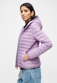 Γυναίκα που φοράει μωβ καπιτονέ μπουφάν με κουκούλα και χέρι στην τσέπη, συνδυασμένο με ανοιχτόχρωμο μπλε τζιν, σε άσπρο φόντο.