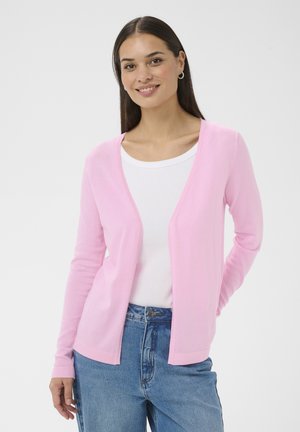 Mosolygó nő halvány rózsaszín kardigánban, fehér felső és kék farmernadrág fölött, egyszerű fehér háttér előtt állva.