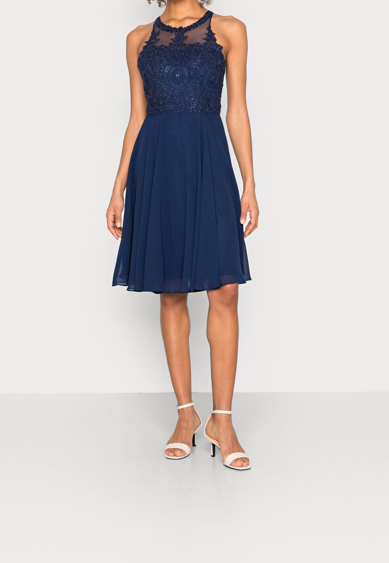 Femme portant une robe bleu marine sans manches, longueur au genou, avec un corsage en dentelle perlée et un décolleté transparent, assortie à des talons blancs à bride cheville.