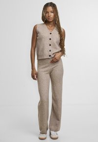 Bež pleteni dvo-dijelni set koji uključuje prsluk bez rukava s kopčanjem na gumbe i V-izrezom te štikle od iste tkanine visokog struka i suženih nogavica. Glatka tekstura, ležeran dizajn.