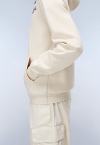 Felpa beige con tasca laterale, polsini e orlo a coste, e un piccolo logo sul petto. Tessuto liscio con una consistenza morbida.