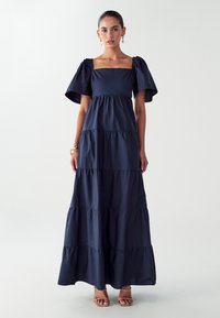 Námořnické maxi šaty se čtvercovým výstřihem, krátkými nabíranými rukávy, vrstvenými volány a hladkou texturou. Nosit s hnědými vysokými podpatky.