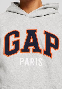 Sudadera con capucha gris con grandes letras bordadas en azul marino y naranja que forman la palabra "GAP" encima de la palabra "PARIS" en blanco.