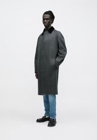 Grå frakke med krave og knaplukning, med sidelommer. Kombineret med blå jeans og sorte sko.