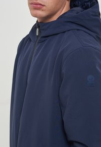 Giacca con cappuccio di colore blu navy realizzata in tessuto resistente all'acqua, dotata di chiusura con zip e dettaglio del logo sulla spalla sinistra. Texture liscia.