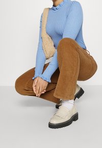 Osoba v podřepu, oblečená v světle modrém žebrovaném svetru, hnědých kordových kalhotách, krémových loaferkách s bílými ponožkami a béžové texturované kabelce na rameno.