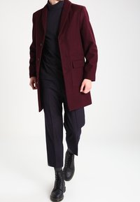 Bordeauxroter Wollmantel mit Reverskragen, einreihig, zwei Fronttaschen, kombiniert mit schwarzem Rollkragenpullover und eng anliegenden dunklen Hosen.