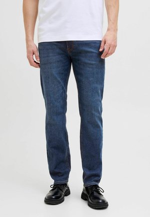 Man draagt blauwe jeans met rechte pijpen, zwarte leren schoenen en een wit overhemd, staand tegen een effen achtergrond.