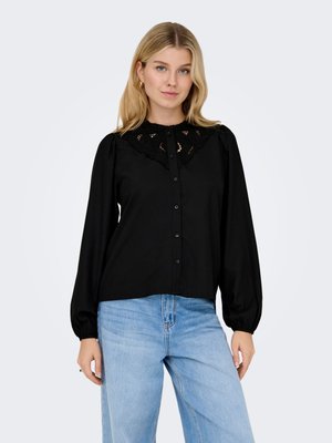 Femme blonde portant un chemisier noir avec des détails en dentelle sur le col et un jean bleu clair taille haute, debout devant un fond uni.