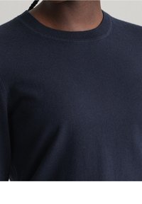 Pull tricoté bleu marine avec un col ras du cou, présentant une texture lisse, un col côtelé et des manches longues. Design minimaliste sans motifs.