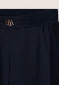 Pantaloni blu navy con cintura in velluto, gamba piatta, tasche laterali e dettaglio del logo metallico in vita. Texture liscia complessiva.
