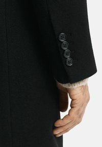 Schwarzer Wollmantelärmel mit vier schwarzen Knöpfen an der Manschette, darunter ein strukturiertes, helles Strickstück. Hand ruht an dem Mantel.