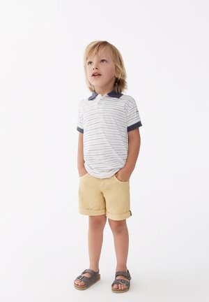 Polo de manga corta a rayas en blanco y azul marino, combinado con pantalones cortos amarillos enrollados y sandalias grises con hebillas.